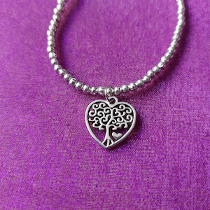 May include: Silver beaded bracelet with a heart-shaped charm. The charm features a tree design with a small heart at the base. The bracelet is displayed on a purple background.