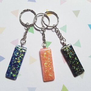 May include: Three keychains with rectangular resin charms. The charms are filled with iridescent glitter and have a blue, pink, and black background. The keychains are attached to silver key rings.
