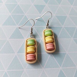 Könnte beinhalten: Ein Paar Hängeohrringe mit einem Stapel von drei pastellfarbenen Macarons auf einem beigefarbenen Hintergrund. Die Macarons sind grün, gelb und rosa.