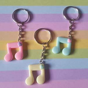 Könnte beinhalten: Drei pastellfarbene Musiknotenschlüsselanhänger mit silbernen Schlüsselringen. Die Schlüsselanhänger sind rosa, hellblau und gelb. Jede Note hat einen kleinen herzförmigen Akzent.