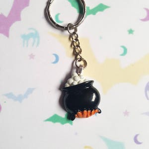 May include: A black cauldron keychain with white bubbling potion and orange flames. The keychain is attached to a silver key ring and chain.