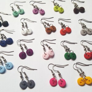 Könnte beinhalten: Sortiment an bunten Knopf-Ohrringen. Die Ohrringe bestehen aus kleinen, runden Knöpfen in verschiedenen Farben, darunter Blau, Grün, Rot, Gelb, Lila, Rosa, Weiß, Schwarz und Grau. Die Knöpfe sind an silbernen Ohrhaken befestigt.
