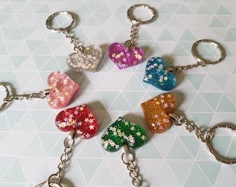 May include: Seven heart-shaped keychains with glitter and star accents. The keychains are in various colors, including pink, red, green, blue, silver, gold, and purple.