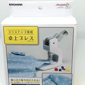 May include: White plastic snap press machine with a blue and white polka dot fabric sample. The machine is labeled "Plus Snap Dedicated Tabletop Press" and "SUN15-94".