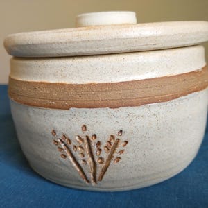 May include: A round, off-white ceramic jar with a matching lid. The jar features a speckled texture, a horizontal brown band around the upper section, and a carved wheat stalk design on the front. The lid has a small, raised central knob.