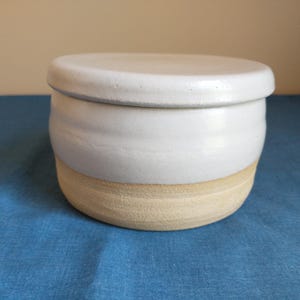 May include: A round ceramic container with a lid, featuring a smooth white glazed upper section and a textured, unglazed beige base. The vessel has a simple, cylindrical shape with a slightly rounded profile and a flat, circular lid on top.