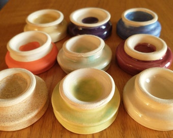 May include: Ten ceramic ring holders in various colors, including white, beige, light blue, light green, orange, purple, and a light blue with a purple glaze. The holders are all round and have a raised lip around the edge.
