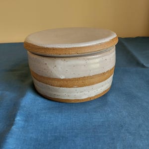 May include: A handmade ceramic butter dish with a lid. The dish is cylindrical with a white and tan striped design. The lid is a matching light gray color. The dish is sitting on a blue surface.