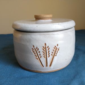 May include: A cylindrical ceramic jar with a white glazed finish and a matching lid. The jar features a carved design of three wheat stalks on the front, revealing the natural tan clay underneath. The lid has a small, raised tan knob handle.