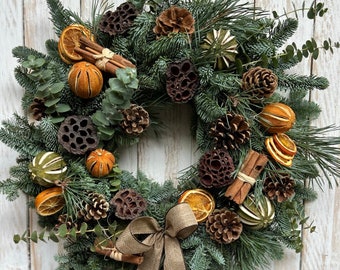 May include: A festive Christmas wreath with a natural aesthetic. The wreath features evergreen branches, pine cones, dried orange slices, cinnamon sticks, and a burlap bow. The wreath is displayed against a white wooden background.