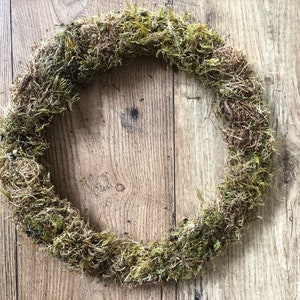 Pu&ograve; includere: Una ghirlanda circolare fatta di muschio naturale, che mostra varie tonalit&agrave; di verde e marrone. Il muschio &egrave; densamente compatto, creando una superficie strutturata. La ghirlanda &egrave; esposta su una superficie di legno con venature visibili.