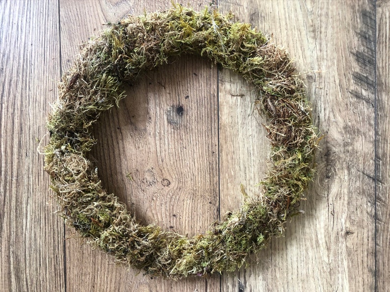 Pu&ograve; includere: Una ghirlanda circolare fatta di muschio naturale, che mostra varie tonalit&agrave; di verde e marrone. Il muschio &egrave; densamente compatto, creando una superficie strutturata. La ghirlanda &egrave; esposta su una superficie di legno con venature visibili.