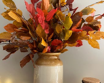 Può includere: Un alto vaso cilindrico in ceramica con la parte superiore color marrone chiaro e la base color crema contiene una grande composizione di foglie dai colori autunnali nelle tonalità del rosso, arancione e giallo. Piccole zucche rosse, pigne marroni e luci circondano la base.