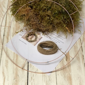 Pu&ograve; includere: Un telaio per ghirlanda in metallo color rame con un mucchio di muschio verde, un rotolo di spago e istruzioni per la creazione di ghirlande. Le istruzioni includono il testo "Happy wreath making". Gli oggetti sono su una superficie di legno bianco.