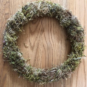 Pu&ograve; includere: Una ghirlanda circolare fatta di muschio verde secco. La ghirlanda &egrave; di colore verde naturale e ha un aspetto ruvido e strutturato. La ghirlanda &egrave; esposta su una superficie di legno.