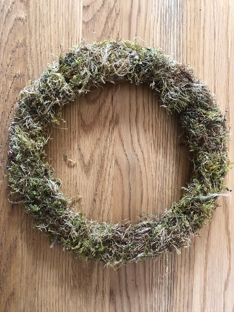 Pu&ograve; includere: Una ghirlanda circolare fatta di muschio verde secco. La ghirlanda &egrave; di colore verde naturale e ha un aspetto ruvido e strutturato. La ghirlanda &egrave; esposta su una superficie di legno.