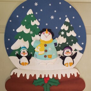 May include: A felt snowman with a blue jacket and yellow scarf stands in a snowy scene with two penguins and two green trees. The scene is surrounded by a blue circle with white snowflakes. The base of the scene is brown with a holly sprig.