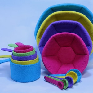 May include: A set of colorful felt measuring cups in shades of blue, green, purple, and pink. The cups are nested together and have a scalloped edge.