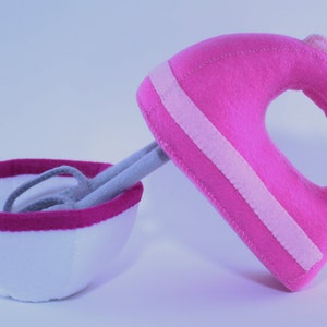May include: A pink felt hand mixer with a white bowl. The mixer has a white stripe on it.