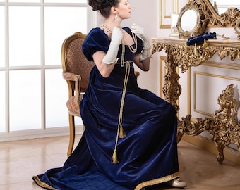 Op de afbeelding: Een vrouw in een blauwe fluwelen jurk met gouden versiering zit op een stoel en past een parelketting aan. Ze draagt witte handschoenen en bevindt zich in een kamer met een grote, sierlijke spiegel en een vergulde kaptafel.