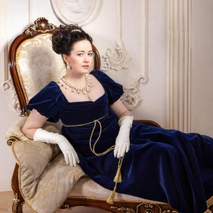 Pu&ograve; includere: Una donna con un abito di velluto blu con una cintura dorata e una collana di perle &egrave; seduta su una chaise longue con guanti bianchi.