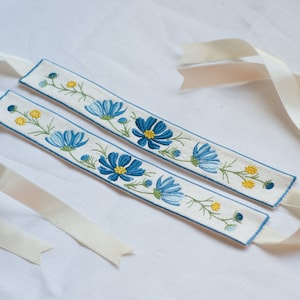 May include: Two white fabric ribbon bookmarks with blue and yellow embroidered flowers. The bookmarks have white satin ribbon ties.