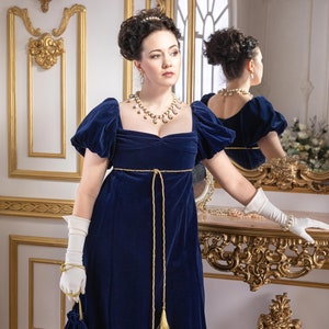 Pu&ograve; includere: Una donna con un abito di velluto blu con una cintura dorata e una collana di perle si trova davanti a uno specchio. Indossa guanti bianchi e tiene una piccola borsa di velluto blu con nappe dorate.