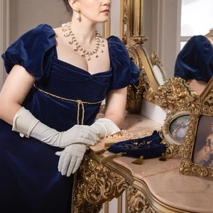 Pu&ograve; includere: Una donna con un abito di velluto blu, una collana e orecchini di perle, si trova di fronte a uno specchio dorato. Indossa guanti bianchi e guarda il suo riflesso. Ci sono due cornici dorate ornate sul tavolo di fronte a lei.