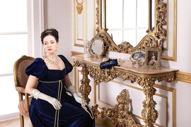Pu&ograve; includere: Una donna con un abito di velluto blu e una collana di perle &egrave; seduta su una sedia di fronte a un tavolo da toeletta con uno specchio dorato e dettagli decorativi. Il tavolo ha un piano in marmo ed &egrave; decorato con una piccola cornice e una pochette in velluto blu.