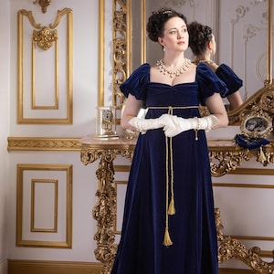 Pu&ograve; includere: Una donna con un abito di velluto blu con rifiniture dorate e una collana di perle si trova in una stanza con accenti dorati e uno specchio.