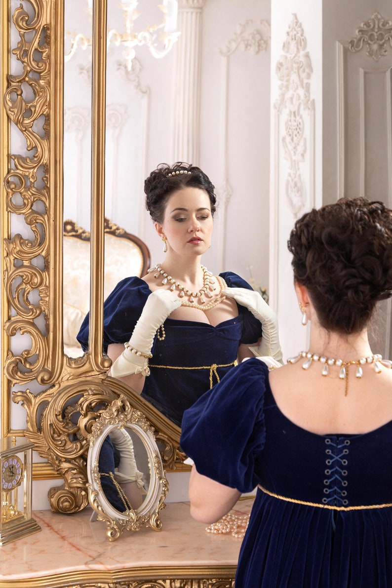 Pu&ograve; includere: Una donna con un abito di velluto blu, una collana di perle e orecchini abbinati, si trova davanti a uno specchio, regolando la sua collana. Lo specchio ha una cornice dorata decorata.