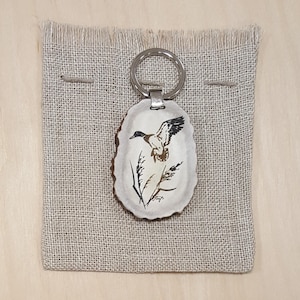 May include: A brown antler keychain with a painted image of a duck in flight. The antler is oval shaped and has a silver key ring attached.