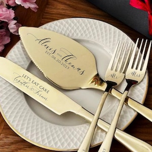 May include: A gold-colored wedding cake knife and server set with the names "Alexis & Thomas" and the date "06.20.2025" engraved. The knife reads "Let's Eat Cake Together & Fermer". Two forks are engraved with "Mr" and "Mrs".