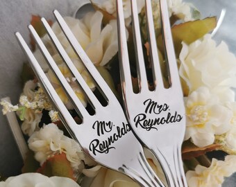 以下が含まれることがあります： 「Mr. Reynolds」と「Mrs. Reynolds」と刻まれた2本の銀のフォークが、白い花の上に置かれています。