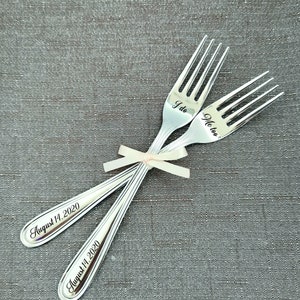 May include: Two silver forks tied together with a white ribbon. The forks are engraved with the text "I do" and "Me too" and the date "August 11, 2020".