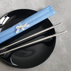 May include: A pair of silver chopsticks with the inscription "GELI &hearts; NORBERT" on each chopstick. The chopsticks are wrapped in a light blue tissue paper with a white ribbon and a small heart charm.