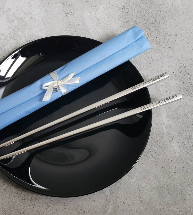 May include: A pair of silver chopsticks with the inscription "GELI &hearts; NORBERT" on each chopstick. The chopsticks are wrapped in a light blue tissue paper with a white ribbon and a small heart charm.