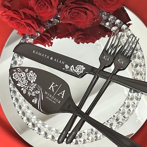 May include: A black wedding cake knife and server set with floral designs and personalized text. Two forks are also included. The set is displayed on a mirrored plate with crystal accents, alongside red roses.