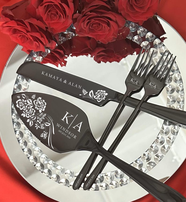 May include: A black wedding cake knife and server set with floral designs and personalized text. Two forks are also included. The set is displayed on a mirrored plate with crystal accents, alongside red roses.