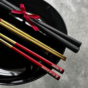 Puede incluir: Un plato negro de cer&aacute;mica contiene palillos chinos negros y dorados, con una cinta roja. Dos palillos chinos rojos est&aacute;n grabados con "MARABEL QUEEN OF MY LIFE" y un s&iacute;mbolo de corona. Juego de regalo de palillos.