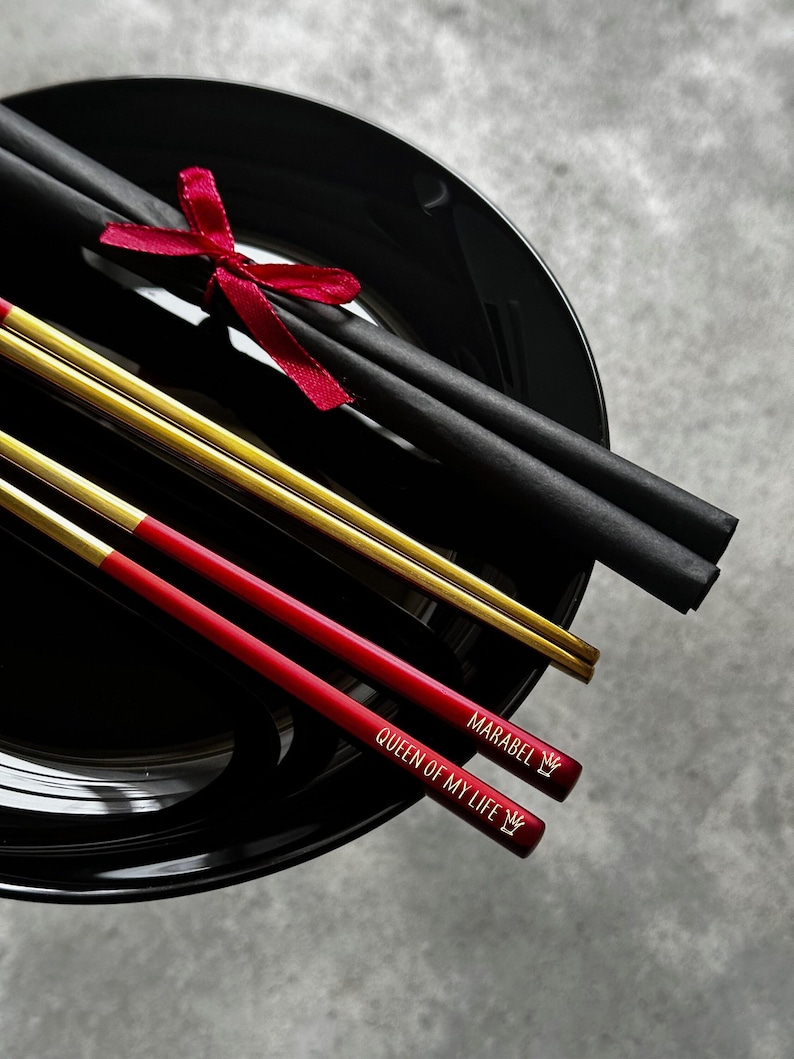 Puede incluir: Un plato negro de cer&aacute;mica contiene palillos chinos negros y dorados, con una cinta roja. Dos palillos chinos rojos est&aacute;n grabados con "MARABEL QUEEN OF MY LIFE" y un s&iacute;mbolo de corona. Juego de regalo de palillos.