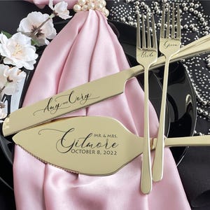 May include: A gold-colored wedding cake knife and server set with the names "Amy and Cory" and "Mr. & Mrs. Gilmore October 8, 2022" engraved. Two forks are also visible, one labeled "Bride" and the other "Groom". The set rests on a pink cloth.