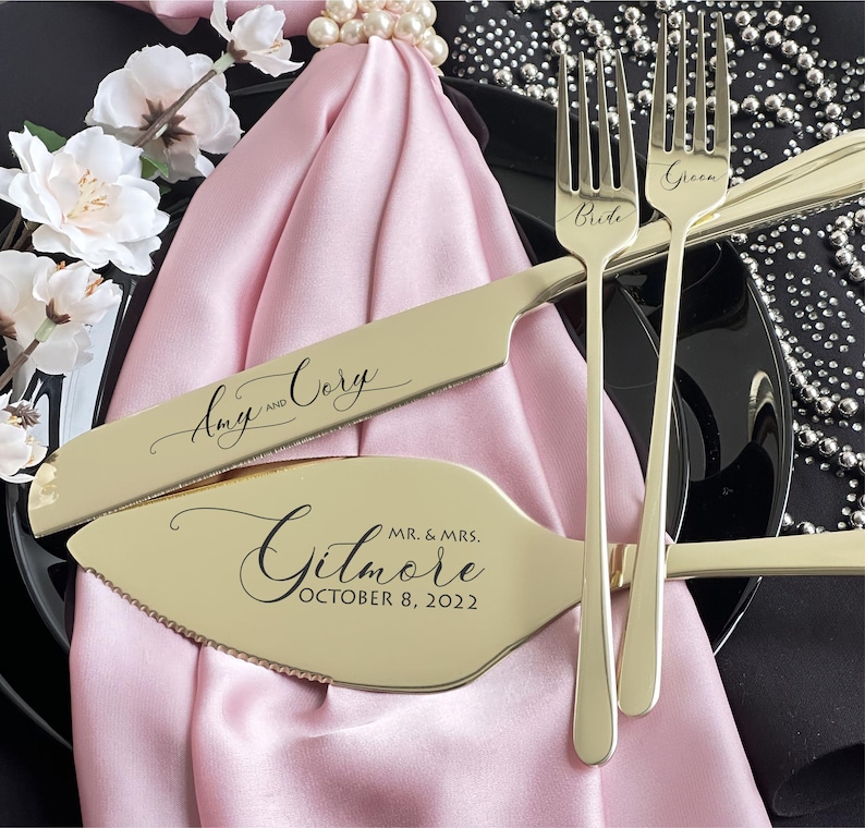 May include: A gold-colored wedding cake knife and server set with the names "Amy and Cory" and "Mr. & Mrs. Gilmore October 8, 2022" engraved. Two forks are also visible, one labeled "Bride" and the other "Groom". The set rests on a pink cloth.