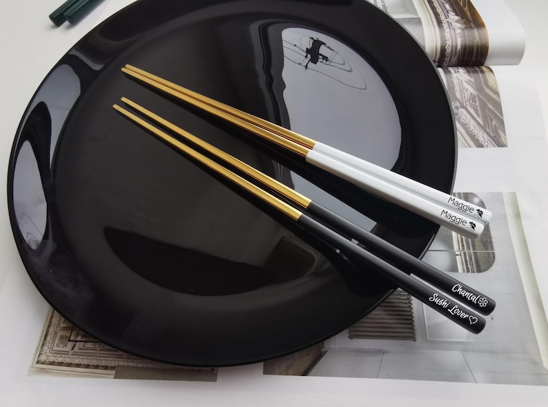 Puede incluir: Dos pares de palillos con puntas doradas, sobre un plato negro. Un par es negro con el texto blanco "Chantal Sushi Lover". El otro par es blanco con el texto "Maggie".