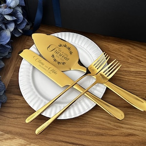 May include: A gold-colored wedding cake serving set and forks are displayed on a white plate. The set includes a cake server and knife, both engraved with names and a date. The forks are also engraved. The set is on a wooden surface.