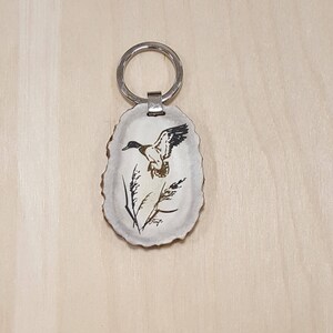 May include: A white antler keychain with a black and brown duck flying over a stalk of grass. The keychain has a silver ring.