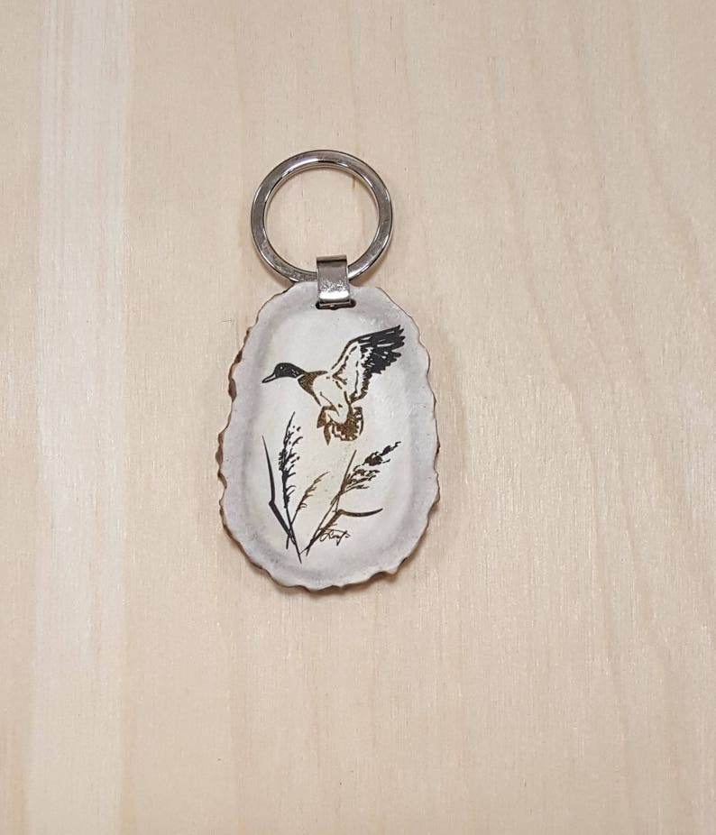 May include: A white antler keychain with a black and brown duck flying over a stalk of grass. The keychain has a silver ring.