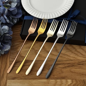 May include: Five forks in various metallic finishes: copper, gold, silver, and black. The forks are arranged on a wooden surface near a white plate with a decorative rim. A black box with a blue ribbon is in the background.
