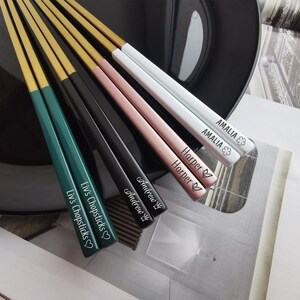 May include: Set of six personalized chopsticks with gold tips. The chopsticks are in different colors: green, black, pink, and white. Each chopstick is engraved with a name: Liv's Chopsticks, Andrew, Amelia, and Harper.
