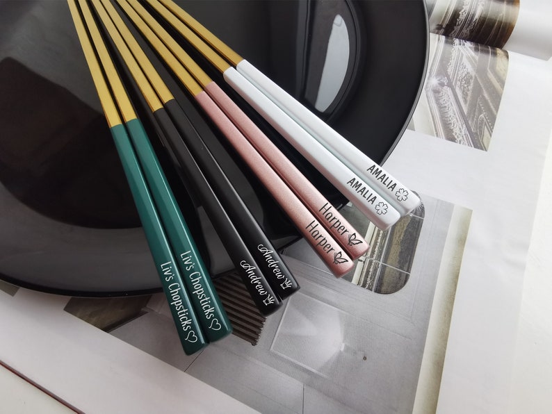 May include: Set of six personalized chopsticks with gold tips. The chopsticks are green, black, pink, and white. The names "Liv's Chopsticks", "Andrew", "Andrew", "Harper", "Amalia", and "Amalia" are engraved on the chopsticks.